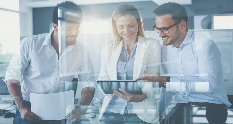 Three business professionals, two men and one woman, are smiling and looking at a tablet together in a bright modern office setting. One man holds papers, and the group appears to be collaborating. EFESO Management Consultants